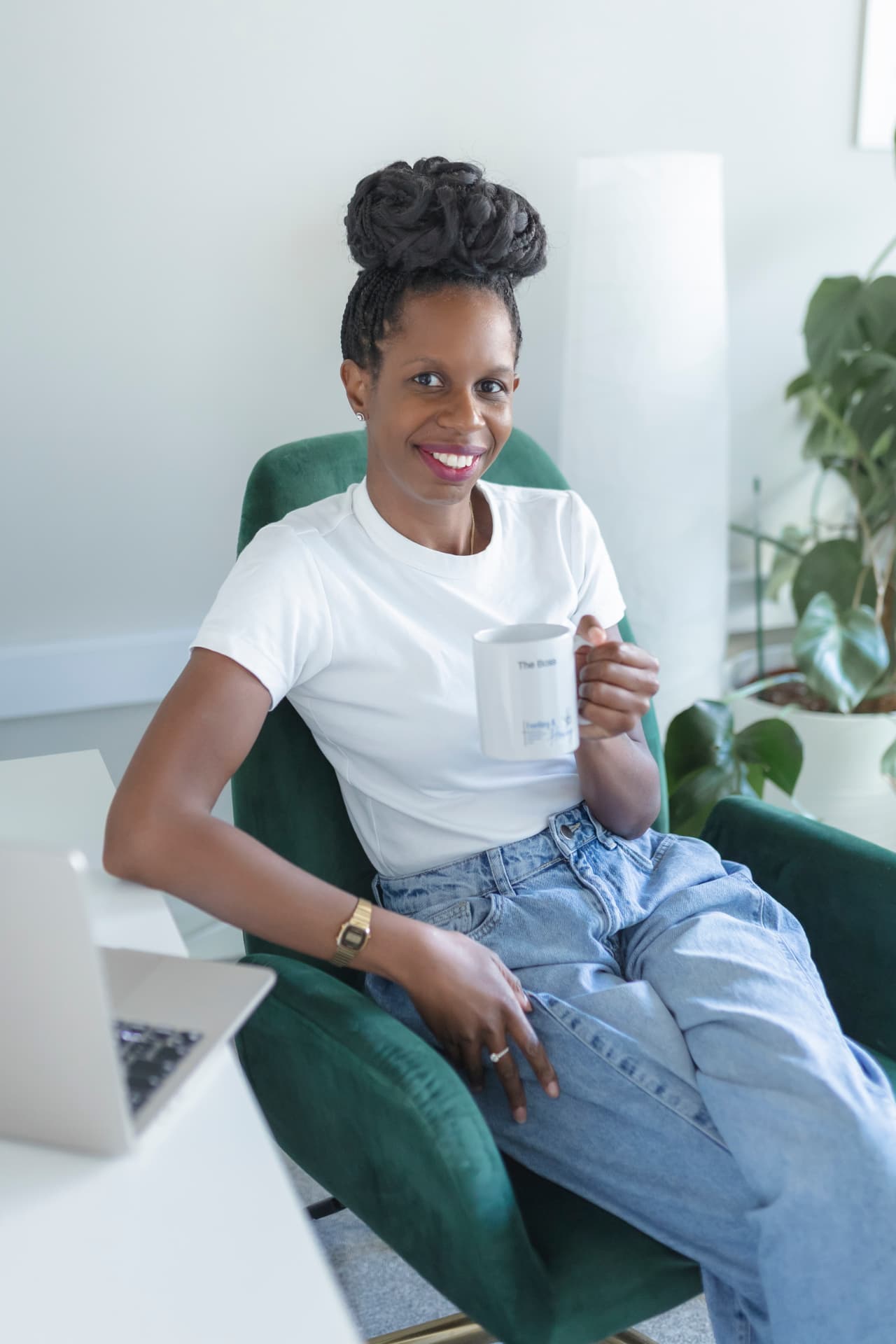 A woman seated in a green armchair beside a desk with an open laptop, holding a mug, in a calm, plant-filled room